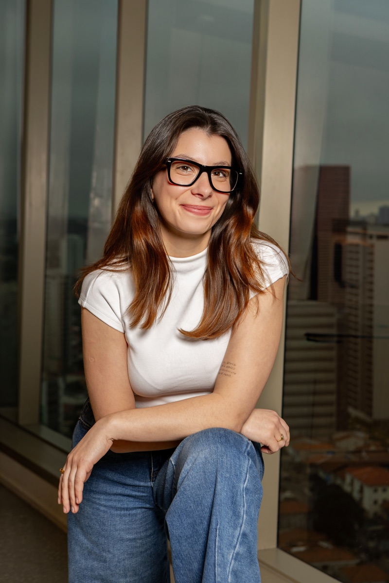 Victoria Giroto sorrindo, de óculos e camiseta branca, sentada perto de uma janela com vista para a cidade